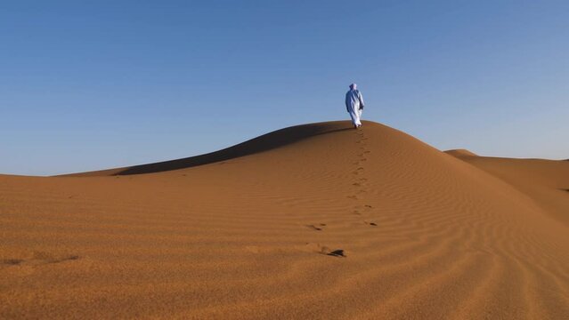 A traditionally dressed Moroccan man wearing a blue gandoura, djellaba and a turban walks on a sand dune, Erg Chegaga desert, Morocco. Authentic desert scene of South Morocco. 4k.