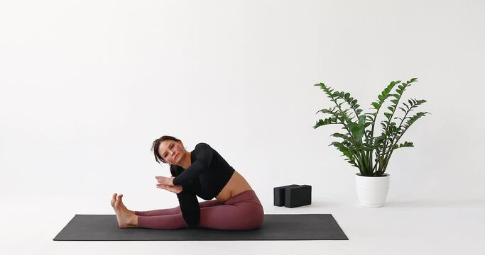 A woman practicing yoga performs the Parivritta Paschimottanasana exercise, twisted side angle pose, training in sportswear while sitting on a mat