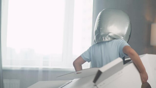 Boy in an astronaut helmet plays with a cardboard space shuttle, runs around the living room, dreamer.