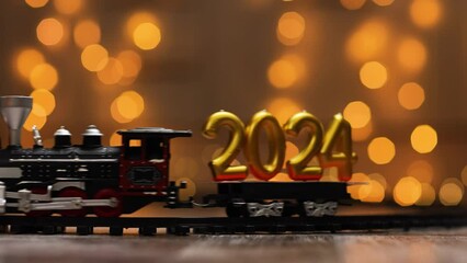 The train delivers the golden figures of the upcoming new year as cargo. Golden Inscription of the upcoming new year 2024 on a background of twinkling lights.