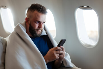 man on the plane. a male passenger sits on an airplane covered with a blanket and holds a phone in his hands. convenience and comfort during the flight concept - obrazy, fototapety, plakaty