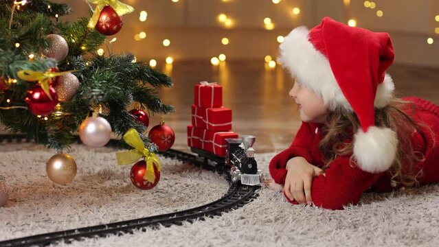 A girl in a Santa hat and a red sweater enthusiastically looks up at a festively decorated Christmas tree and decorations. A toy fairy - tale train brings Christmas gifts to children . Background of l