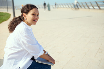 Young freelance entrepreneur woman holding laptop while resting outdoors after work, smiling broadly looking aside