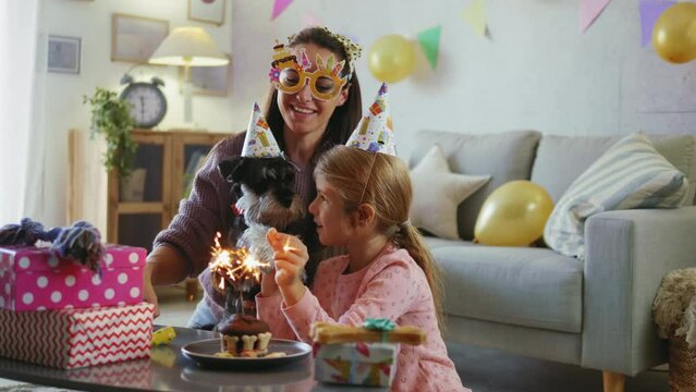 Mother and daughter celebrate their dog's birthday with birthday hats and cake, singing, and taking selfies or videos for social media at home