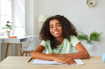 Webcam portrait of smiling dark-skinned schoolgirl who having online class learning at home. Joyful cute curly african american teenage girl with notebook sitting at table in front of web camera.