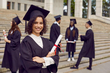 Young smiling happy girl student in a university graduate gown and diploma in her hands. Woman with crossed arms outdoor on the background of classmates. Graduation and education concept.