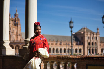 Obraz premium Young black and South American woman in a beige gypsy flamenco suit, posing in a beautiful square in the city of Seville in Spain. Concept dance, folklore, flamenco, art.