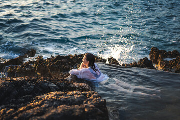 Young cheerful relaxed woman in a white shirt in a natural sea bath on the seashore. Thalassotherapy wellness concept