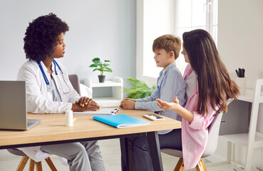 Obraz premium Female pediatrician talking to small boy patient and his mother in medical office. African American doctor, general practitioner doing medical checkup of little patient in clinic. Children healthcare