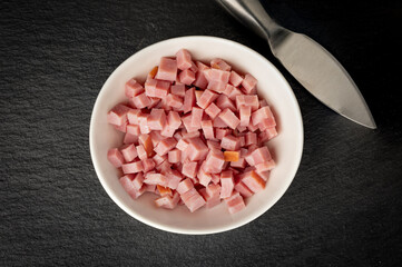 A cup with diced ham placed on black stone background next to small kitchen knife
