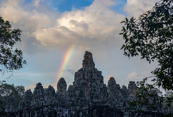 As the sun sets.  Bayon temple, Siem Reap, Cambodia in the late evening light.  A rainbow is...
