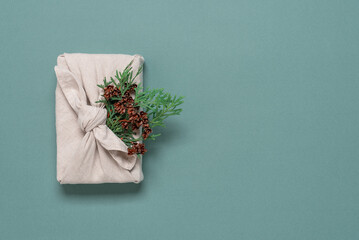 Christmas gift wrapped in fabric with conifer branch, green pastel background. A traditional furoshiki gift. Top view,flat lay.