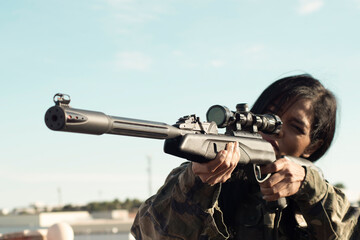 A female sniper with dark skin in camouflage military clothing holds a gun, takes aim, selective...
