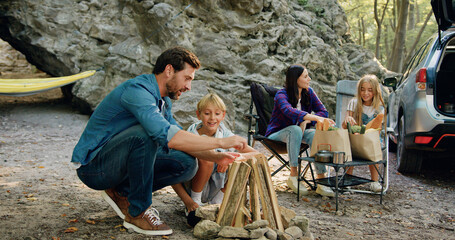 Father and son sitting together make fire at picnic, making firewood, mother and daughter having fun talking in the background. Outbound family trip with a trailer camper to the mountains.