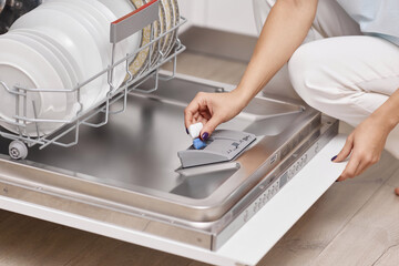 female hand puts dishwasher tablet into open automatic built-in dishwasher machine with dirty dish inside in kitchen - obrazy, fototapety, plakaty