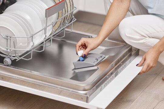 female hand puts dishwasher tablet into open automatic built-in dishwasher machine with dirty dish inside in kitchen