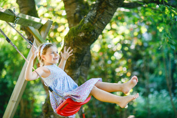 Happy little preschool girl having fun on swing in domestic garden. Cute healthy child swinging under blooming trees on sunny spring day. Kid laughing and crying.