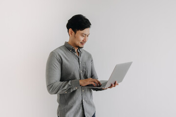 Asian man with beard wear grey shirt typing keyboard working on notebook and holding laptop, happy smiling face looking at computer and standing isolated over white background wall.