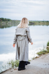 Sad girl walking along the embankment of the river, back view of a girl in a raincoat, autumn clothes. Blonde woman on the lake.