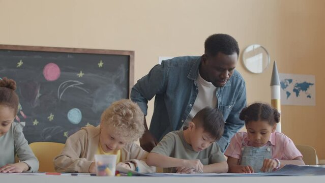 African American teacher helping multi-ethnic boys and girls to draw pictures with wax crayons while giving art class in primary school