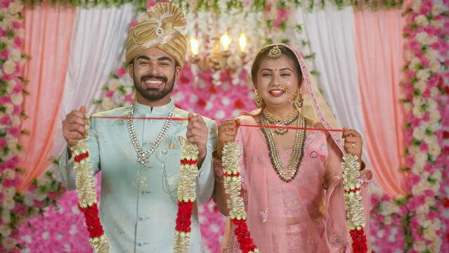 happy smiling newlywed couples showing garlands by looking at camera on weeding or marriage reception stage - concept of ceremony, traditional culture and fashion wear.