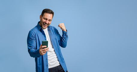 Successful male entrepreneur pumping fist after reading good news over cellphone on blue background - obrazy, fototapety, plakaty
