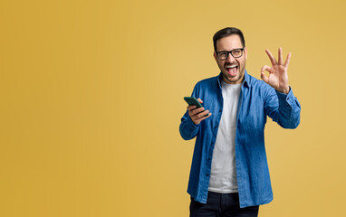 Handsome freelancer shouting happily and showing OK sign while using cellphone on yellow background - obrazy, fototapety, plakaty