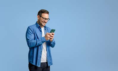 Smiling handsome businessman text messaging over mobile phone while standing on blue background - obrazy, fototapety, plakaty