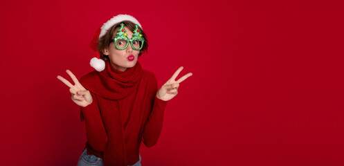 Excited beautiful smiling young woman in Santa hat and Christmas glasses is posing and having fun on party on red background. Merry christmas. Happy new year. Holidays and celebration in winter