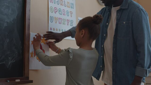 African American male teacher helping little girl to hang space rocket drawing on wall in classroom
