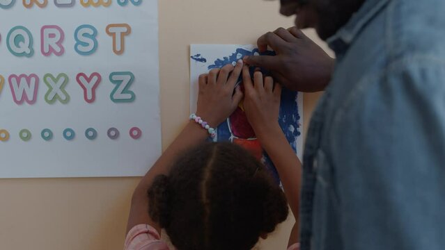 Follow shot of little Black girl walking in classroom and putting paper drawing of space rocket on wall with help of male teacher