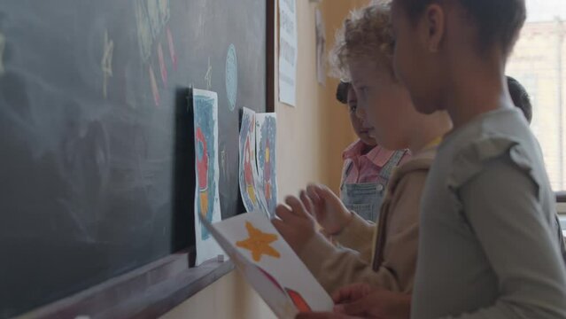 Group of little multi-ethnic kids placing paper drawings of space rockets by chalkboard while preparing art exhibition in classroom