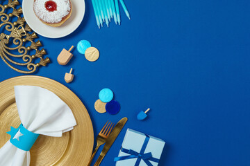 Hanukkah table setting concept with plate, stylish cutlery, gift box and traditional donuts on blue background. Top view, flat lay