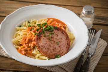 Jägerschnitzel nach DDR GDR Rezept mit Jagdwurst gebraten, Makkaroni Penne Nudeln, Tomatensoße, Petersilie mit Porzellan tiefer Teller, Besteck, Salzstreuer, Serviette, rustikal dunkel Holz Hintergund