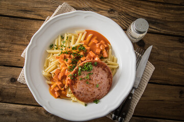 Jägerschnitzel nach DDR GDR Rezept mit Jagdwurst gebraten, Makkaroni Penne Nudeln, Tomatensoße, Petersilie mit Porzellan tiefer Teller, Besteck, Salzstreuer, Serviette, rustikal dunkel Holz Hintergund