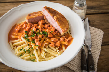 Jägerschnitzel nach DDR GDR Rezept mit Jagdwurst paniert, Makkaroni Penne Nudeln, Tomatensoße, Petersilie mit Porzellan tiefer Teller, Besteck, Salzstreuer, Serviette, rustikal dunkel Holz Hintergund