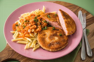 Jägerschnitzel nach DDR GDR Rezept mit Jagdwurst paniert, Makkaroni Penne Nudeln, Tomatensoße, Petersilie mit pink rosa Teller, Besteck, Holzbrett, Serviette und grünen Hintergrund