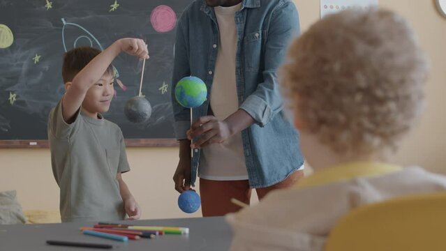 Little Asian boy holding planet model and answering questions of Black teacher while standing in front of chalkboard in classroom durian astronomy lesson