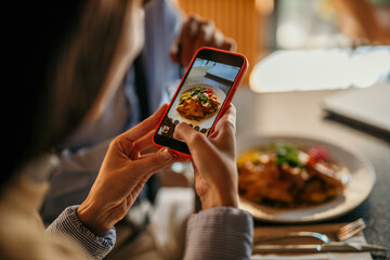 Female taking a photo with a smartphone of a food before eating in the bright venue. - obrazy, fototapety, plakaty
