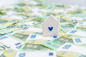 A blue heart on a little white house placed on many euro money bills, concept of costly owning a house - obrazy, fototapety, plakaty