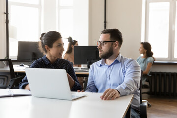 Diverse business colleagues cooperating on project tasks at laptop, sitting at work desk, talking,...