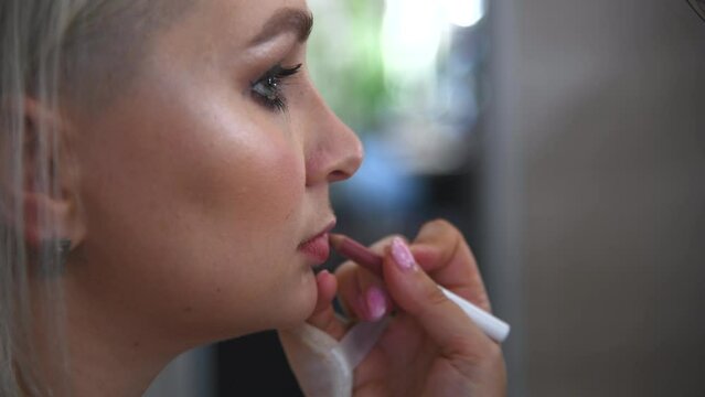 Close-up view of make-up artist applying lipstick using pink wax pencil on lips of adult blond haired woman in beauty salon. Soft focus. Real time handheld video. Make-up and beauty industry theme.