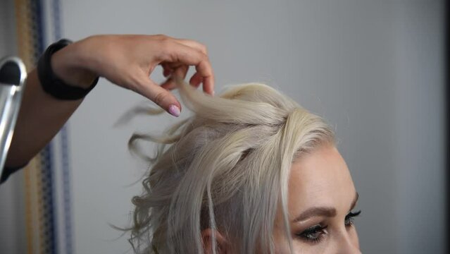 Close-up view of hairdresser making new coiffure for adult blond haired woman using curling iron and comb in hair salon. Soft focus. Real time handheld video. Beauty industry theme.