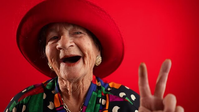 Playful, elderly mature woman, 80s, giving peace sign gesture with no teeth and red hat, isolated on red background studio. Concept of youthful old lady in slow motion