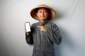Happy Asian farmer wearing Lurik and Caping looking at camera while showing blank mobile phone screen and pointing on it