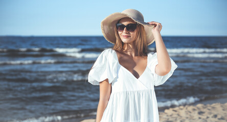 Happy blonde woman is posing on the ocean beach with sunglasses and a hat. Evening sun