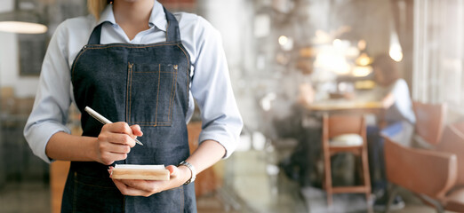 Female waitress wearing apron standing in cozy coffeehouse with notepad in hands to take order in restaurant - Powered by Adobe