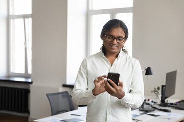Happy Indian businessman in glasses typing on mobile phone at workplace table, reading online chat, text message on smartphone, using Internet service, online app for communication