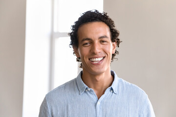 Happy cheerful handsome young Latin businessman closeup head shot portrait. Attractive curly haired man in casual shirt with white teeth standing indoors, looking at camera, smiling