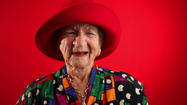Slow motion view of concerned, unhappy elderly senior old funny crazy woman with wrinkled skin red hat and no teeth put hands out ask WHY ME, isolated on red background studio.